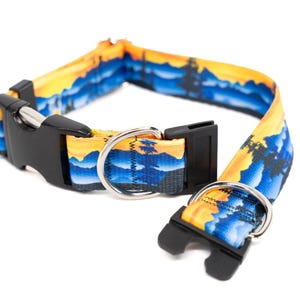 May include: A dog collar with a landscape design featuring a sunset with yellow, orange, and blue hues. The collar has a black plastic buckle and a silver metal D-ring. The collar is made of a durable fabric.