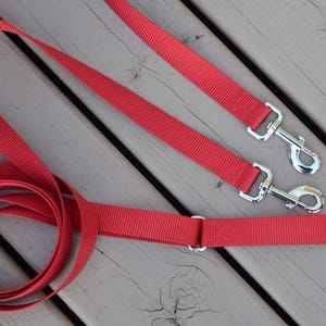 May include: A bright red nylon dog leash featuring a durable woven texture, a sturdy metal swivel snap hook at one end, and a secondary metal swivel snap hook attached to a sliding metal ring, allowing for adjustable length and multiple connection points.