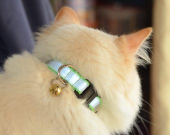 May include: A cat collar featuring a bright lime green nylon strap with a reflective silver strip running down the center. The collar includes a black plastic side-release buckle, a gold-toned metal D-ring, and a small, round gold-colored bell.