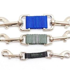 May include: Three double-ended swivel snap hooks connected by short nylon webbing straps. The top strap is bright blue, the middle is olive green, and the bottom is black. Each metal hook features a spring-loaded gate and a polished silver finish.