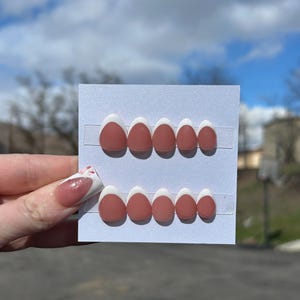 May include: A set of press-on nails featuring a matte, terracotta-colored base with a white French tip design. The nails are displayed on a white card, showcasing various sizes. The image is taken outdoors with a blurred background.