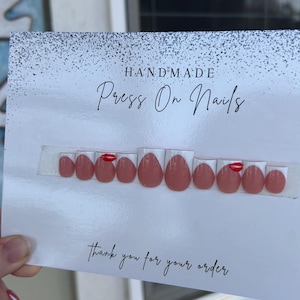 K&ouml;nnte beinhalten: Ein Set handgefertigter Press-On-N&auml;gel in verschiedenen Gr&ouml;&szlig;en, in Nude-Rosa mit einem roten Lippen-Design auf einigen N&auml;geln. Die N&auml;gel werden auf einer Karte mit der Aufschrift "Thank you for your order" pr&auml;sentiert.