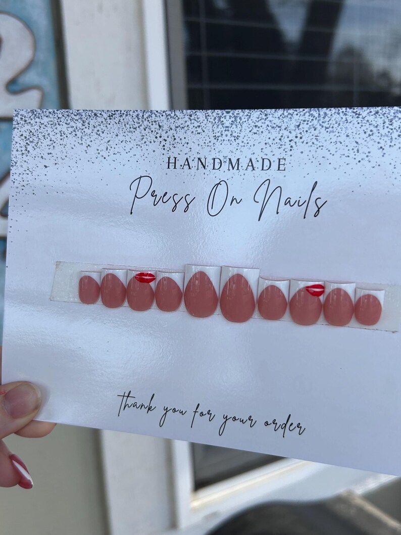 K&ouml;nnte beinhalten: Ein Set handgefertigter Press-On-N&auml;gel in verschiedenen Gr&ouml;&szlig;en, in Nude-Rosa mit einem roten Lippen-Design auf einigen N&auml;geln. Die N&auml;gel werden auf einer Karte mit der Aufschrift "Thank you for your order" pr&auml;sentiert.