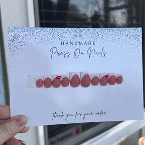 K&ouml;nnte beinhalten: Ein Set handgefertigter Press-On-N&auml;gel in hellem Rosa, mit roten Akzenten. Auf einer wei&szlig;en Karte mit der Aufschrift "Handmade Press On Nails" und "Thank you for your order" pr&auml;sentiert.