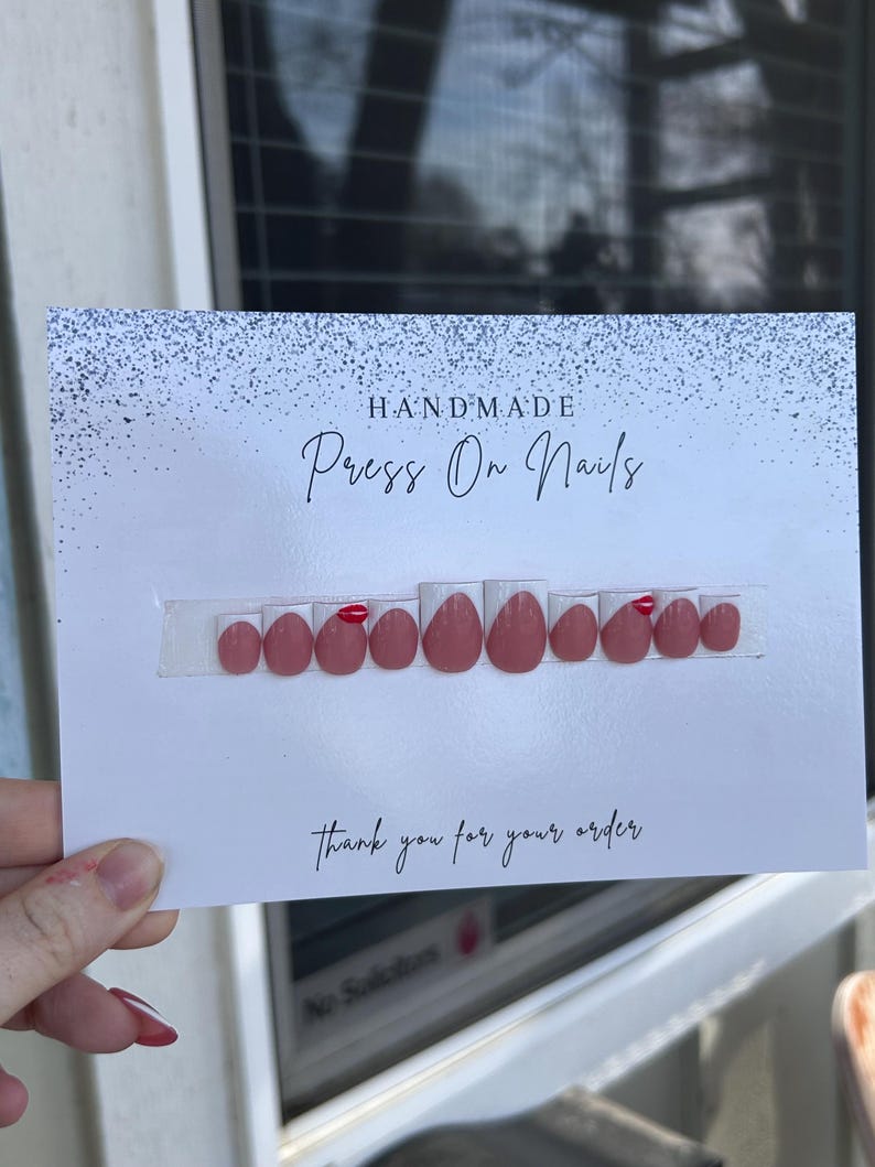 K&ouml;nnte beinhalten: Ein Set handgefertigter Press-On-N&auml;gel in hellem Rosa, mit roten Akzenten. Auf einer wei&szlig;en Karte mit der Aufschrift "Handmade Press On Nails" und "Thank you for your order" pr&auml;sentiert.