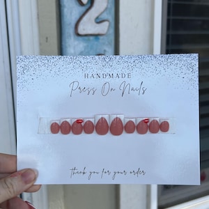 K&ouml;nnte beinhalten: Eine Karte mit handgemachten Press-On-N&auml;geln. Die N&auml;gel sind in einem ged&auml;mpften Rosa gehalten und haben ein gl&auml;nzendes Finish. Einige N&auml;gel haben einen kleinen roten Akzent. Auf der Karte steht "Vielen Dank f&uuml;r Ihre Bestellung" in Schreibschrift.