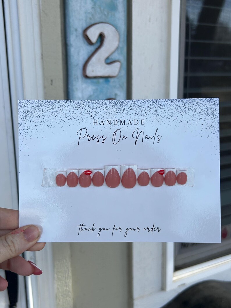 K&ouml;nnte beinhalten: Eine Karte mit handgemachten Press-On-N&auml;geln. Die N&auml;gel sind in einem ged&auml;mpften Rosa gehalten und haben ein gl&auml;nzendes Finish. Einige N&auml;gel haben einen kleinen roten Akzent. Auf der Karte steht "Vielen Dank f&uuml;r Ihre Bestellung" in Schreibschrift.