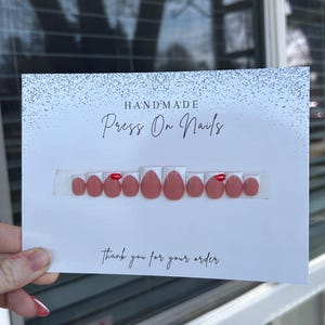 K&ouml;nnte beinhalten: Ein Set handgefertigter Press-On-N&auml;gel in verschiedenen Gr&ouml;&szlig;en, pr&auml;sentiert auf einer wei&szlig;en Karte. Die N&auml;gel sind haupts&auml;chlich in einem ged&auml;mpften Rosa mit gl&auml;nzendem Finish gehalten, einige haben einen roten Akzent. Auf der Karte steht "HANDMADE Press On Nails" und "thank you for your order."
