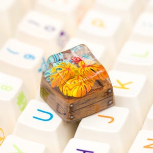 May include: A custom artisan keycap featuring a miniature scene of orange and yellow flowers inside a wooden chest. The keycap is set against a background of white keyboard keys with various symbols and letters.