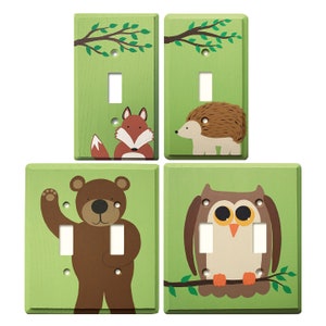 May include: Four light switch covers with a woodland theme. Each cover features a different animal: a fox, a hedgehog, a bear, and an owl. The covers are painted on a green background with brown accents.