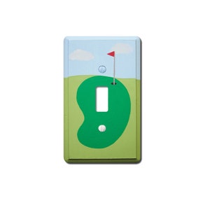 May include: A light switch cover with a green golf putting green design. The putting green has a white golf ball and a red flag in the hole. The background is a light blue sky with white clouds.