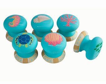 May include: Set of six turquoise blue cabinet knobs with a variety of sea life designs, including a seahorse, seashell, coral, turtle, and seaweed. Each knob has a gold and white base.