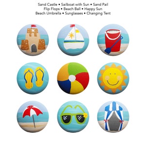 Op de afbeelding: Negen beschilderde ronde stenen met strandthema's. De ontwerpen omvatten een zandkasteel, een zeilboot, een emmer, slippers, een strandbal, de zon, een parasol, een zonnebril en een kleedhok.