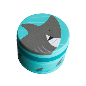 Op de afbeelding: Een tealblauwe houten doos met een geschilderd beeld van een haai met witte tanden. De doos heeft een tealblauwe band rond het midden en kleine haaienvin-ontwerpen rond de bodem.