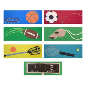 May include: A set of colorful wooden signs with illustrations of different sports, including basketball, soccer, football, lacrosse, tennis, and a whistle. The signs also include a scoreboard with the words "HOME" and "AWAY" and the numbers "3" and "2".
