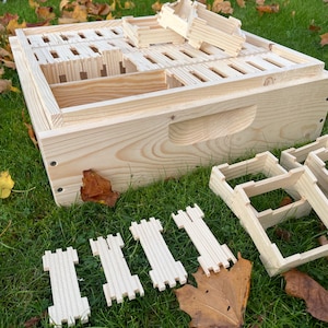 Op de afbeelding: Een gedemonteerde houten bijenkorf met verschillende onderdelen op het gras. De hoofdbehuizing van de korf is rechthoekig met een sleufdeksel en een handvat. Verschillende kleine houten frames en andere onderdelen liggen in de buurt. De natuurlijke houtkleur is zichtbaar.