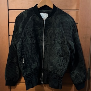 May include: A black bomber jacket with a textured, patterned design. The jacket features a full-length zipper, a ribbed collar, cuffs, and hem. The jacket is hanging on a wooden hanger against a wooden door.