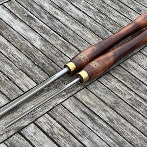 Könnte beinhalten: Zwei Drechselwerkzeuge mit polierten Stahlklingen und dunklen Holzgriffen. Die Griffe haben eine Messingzwinge und ein Logo. Die Werkzeuge liegen auf einer verwitterten Holzoberfläche.