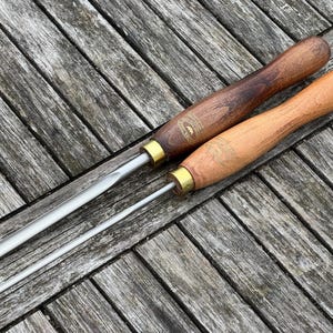 Op de afbeelding: Twee houtdraaigereedschappen met houten handvatten en metalen bladen. Eén handvat is donkerbruin, de andere is lichtbruin. Beide gereedschappen hebben messing hulzen. De gereedschappen liggen op een verweerd houten oppervlak.