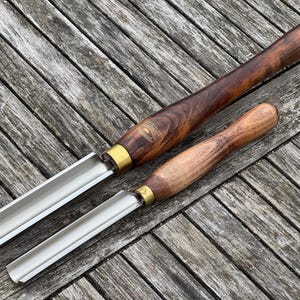 Op de afbeelding: Twee houtdraaiguts met houten handvatten en metalen bladen liggen op een verweerd houten oppervlak. De gutsijzers hebben messing hulzen en zijn verschillend van grootte. De houten handvatten zijn donkerbruin en lichtbruin.