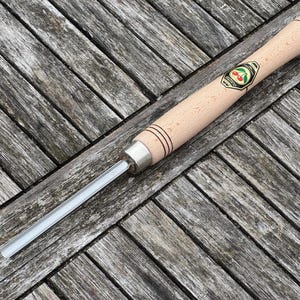 Op de afbeelding: Een houtdraaigereedschap met een lichtgekleurde houten handgreep en een zilveren metalen ferrule. De handgreep heeft een kersenlogo en zwarte ringen. Het lemmet van het gereedschap is recht en zilver. Het gereedschap ligt op een verweerd houten oppervlak.