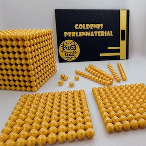 Op de afbeelding: Een verzameling goudgele educatieve wiskundematerialen, waaronder een grote kubus, platte vierkanten, staven en losse kralen. Op de achtergrond een zwarte doos met gouden letters en een logo. De tekst op de doos luidt "GOLDENES PERLENMATERIAL".