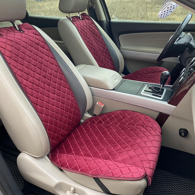May include: Burgundy car seat covers with a diamond quilted pattern. The covers are installed in a car with tan leather seats and a gray and tan interior. The covers are designed to protect the seats and add a touch of style.