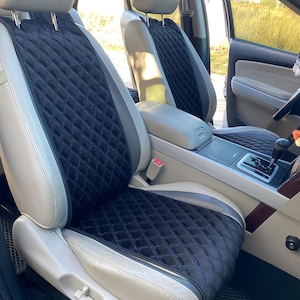 以下が含まれることがあります： 黒いキルティングダイヤモンド模様と青いステッチが施された車のシートカバー。カバーには、ライトグレーと白のアクセントがあります。センターコンソールやステアリングホイールなど、車の内装が見えます。