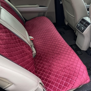 May include: Burgundy car seat cover with a diamond quilted pattern. The cover is made of a soft, velvety material and is designed to fit the back seat of a vehicle. The cover is shown installed in a car with light-colored seats.
