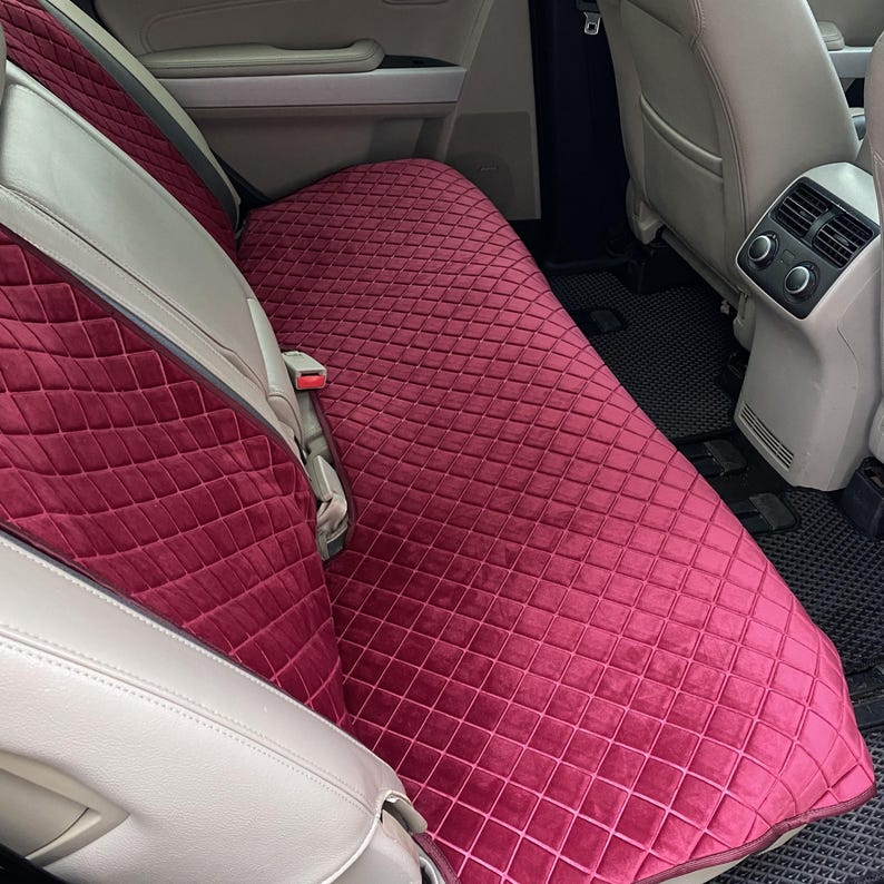 May include: Burgundy car seat cover with a diamond quilted pattern. The cover is made of a soft, velvety material and is designed to fit the back seat of a vehicle. The cover is shown installed in a car with light-colored seats.