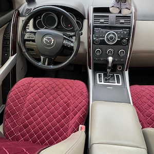 May include: Interior view of a car's front cabin. The steering wheel has a Mazda logo. The dashboard features a radio and climate controls. The seats are covered with burgundy quilted fabric. Small baby shoes rest on the dashboard.