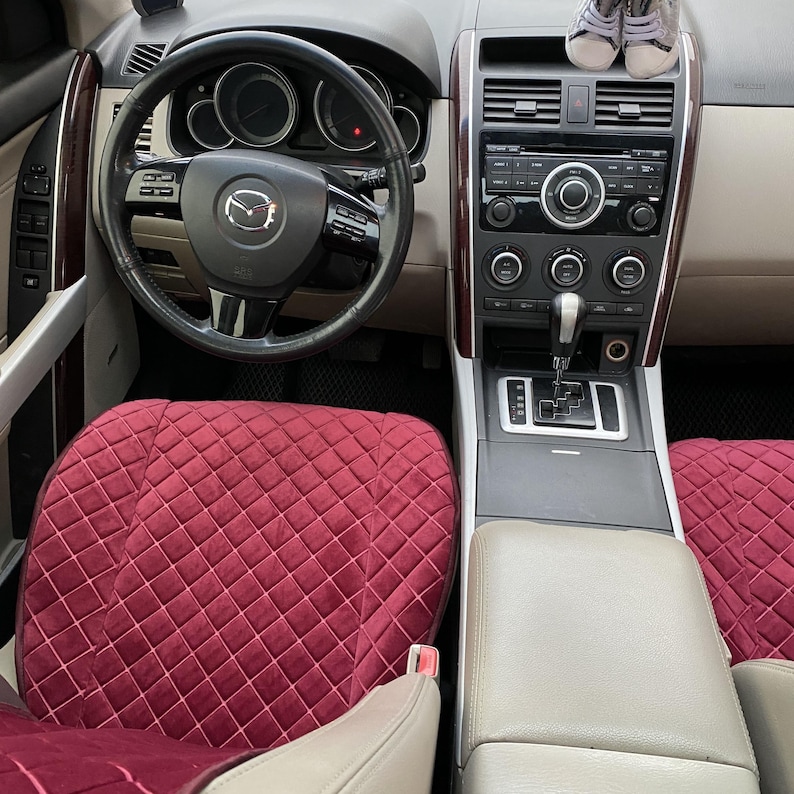 May include: Interior view of a car's front cabin. The steering wheel has a Mazda logo. The dashboard features a radio and climate controls. The seats are covered with burgundy quilted fabric. Small baby shoes rest on the dashboard.