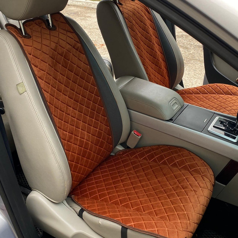 May include: Car seat covers in a vehicle interior. The covers are a warm orange-brown color with a diamond quilted pattern. The seats have a light beige leather trim and dark gray accents. The center console and dashboard are visible.