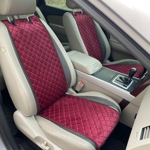 以下が含まれることがあります： ダイヤモンド柄のワインレッドのキルティングされた車のシートカバー。カバーは車のフロントシートに取り付けられています。