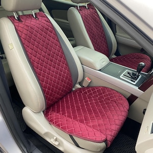 May include: Burgundy car seat covers with a diamond-quilted pattern. The covers are made of a soft, velvety material and are designed to fit over the existing seats. The seats have a light beige leather with gray accents.