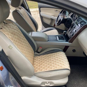 以下が含まれることがあります： ベージュとグレーのキルティングシートカバーが付いた車の内装。シートはダイヤモンドパターンで、黒いトリムがアクセントになっています。ダッシュボードとステアリングホイールが見え、ニュートラルな配色です。車内は明るく照らされています。
