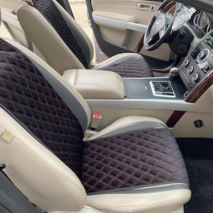 以下が含まれることがあります： ベージュレザーシートと黒いシートカバーを備えた車の内装。シートカバーは、赤いステッチのダイヤモンドキルト模様です。ダッシュボードとステアリングホイールが背景に見えます。