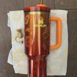 May include: A red and gold Stanley tumbler with a floral design, orange handle, and straw. The tumbler has the Stanley logo in green. A gold pineapple bottle stopper is also present.