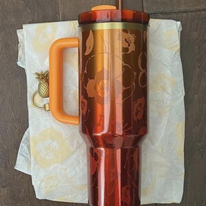 May include: An orange and copper insulated tumbler with a floral pattern. It features a matching orange handle and lid, a brown straw, and a gold pineapple charm. The tumbler is on floral patterned tissue paper.