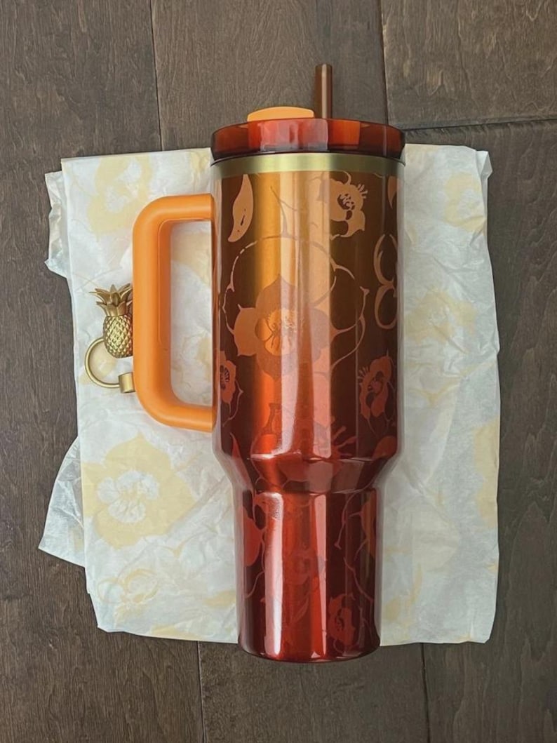 May include: An orange and copper insulated tumbler with a floral pattern. It features a matching orange handle and lid, a brown straw, and a gold pineapple charm. The tumbler is on floral patterned tissue paper.