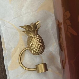 May include: A gold-coloured pineapple-shaped bottle stopper. The stopper has a textured surface and a curved handle that connects to a cylindrical base. The pineapple top has detailed leaves.