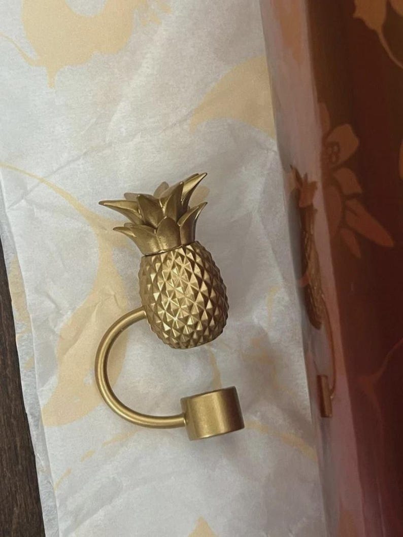 May include: A gold-coloured pineapple-shaped bottle stopper. The stopper has a textured surface and a curved handle that connects to a cylindrical base. The pineapple top has detailed leaves.
