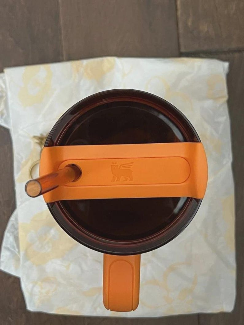May include: Overhead shot of a brown insulated tumbler with an orange handle and lid. The lid features an orange band with a logo and a clear straw. The tumbler is on a white cloth.