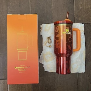 May include: A red and gold Stanley Quencher 112.0 Tumbler with a handle and straw, next to its orange box. The box reads "Quencher 112.0 Tumbler 40 oz (1.18L)". A gold pineapple charm is also present.