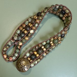 May include: Handmade beaded belt featuring a mix of green, brown, and gold beads. The belt is constructed with a brown leather backing and a decorative metal clasp with a floral design. The beads are various sizes and shapes.