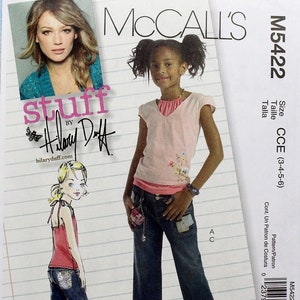 May include: A sewing pattern for a girl's outfit, featuring a pink short-sleeved shirt with a scoop neck and a pair of blue denim capris. The pattern is from McCall's and is size CCE (3-4-5-6).