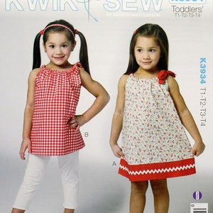 May include: A sewing pattern for toddler dresses and tops. The pattern cover features two young girls modeling the garments. One girl wears a red and white gingham top with white pants. The other wears a floral dress with a red trim.