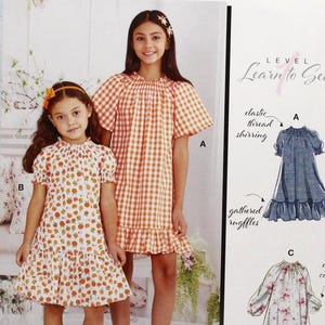May include: McCall's sewing pattern M8283 featuring two young girls wearing dresses. One dress is orange gingham, the other is white with orange floral print. The pattern includes illustrations and the text "Learn to Sew".