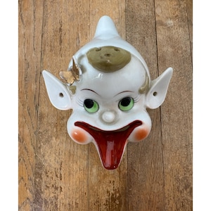 Puede incluir: Máscara de pared de cerámica vintage con una cara de payaso caprichosa. La máscara tiene una base blanca con ojos verdes, una lengua roja y mejillas naranjas. Una mariposa dorada y un sombrero marrón adornan la parte superior. La máscara tiene orejas grandes.