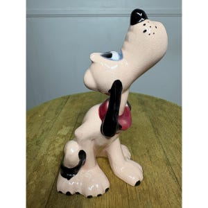 Pode incluir: Uma estatueta de cão em cerâmica com um acabamento brilhante em bege claro, apresentando um nariz preto, orelhas pretas e uma coleira vermelha. O cão está representado numa posição sentada com a cabeça inclinada para cima e olhos azuis.